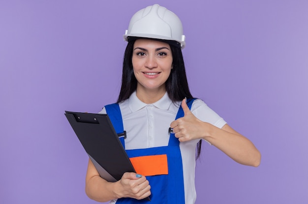 Giovane donna del costruttore in uniforme da costruzione e casco di sicurezza che tiene appunti