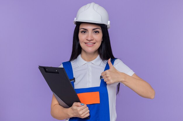 Giovane donna del costruttore in uniforme da costruzione e casco di sicurezza che tiene appunti