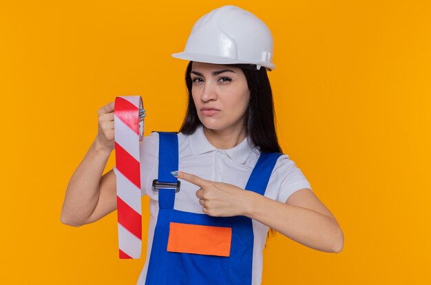 Giovane donna del costruttore in uniforme da costruzione e casco di sicurezza che tiene il nastro adesivo che punta con il dito indice verso di esso guardando davanti con la faccia seria in piedi sopra la parete arancione