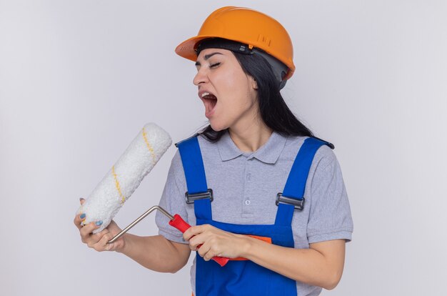 Giovane donna del costruttore in uniforme da costruzione e casco di sicurezza che tiene il rullo di vernice usandolo come microfono che canta divertendosi in piedi sopra il muro bianco