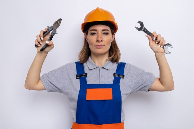 Giovane donna del costruttore in uniforme da costruzione e casco di sicurezza che tiene la chiave e le pinze con un'espressione seria e sicura in piedi sul muro bianco