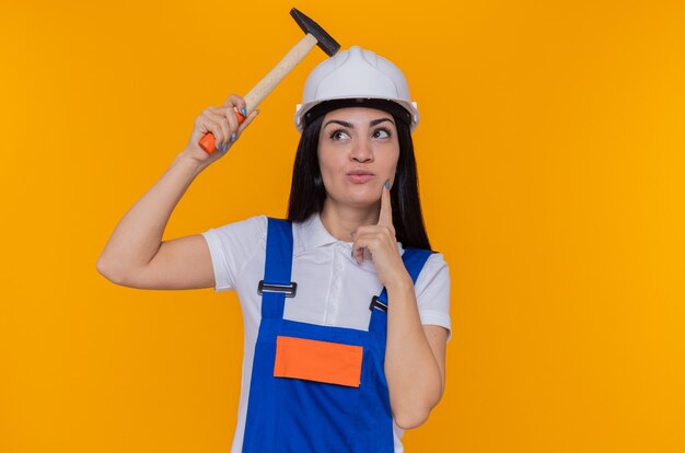 Giovane donna del costruttore in uniforme da costruzione e casco di sicurezza che tiene martello che osserva in su con espressione pensierosa sul pensiero del viso in piedi sopra la parete arancione