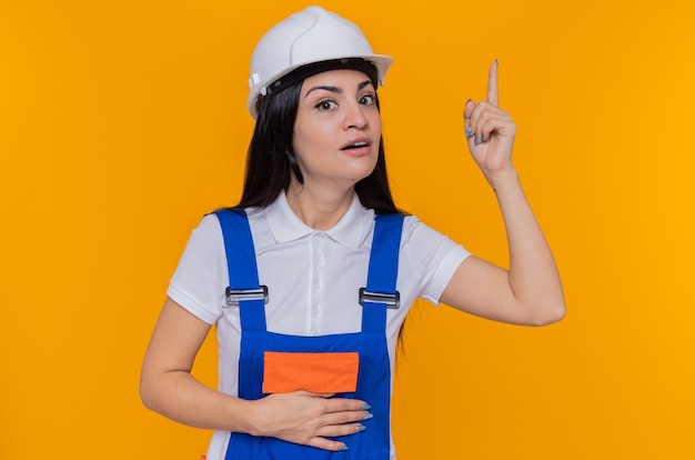 Giovane donna del costruttore in uniforme da costruzione e casco di sicurezza guardando la parte anteriore con il sorriso sul viso sorpreso intelligente che mostra il dito indice che ha una grande idea in piedi sopra la parete arancione