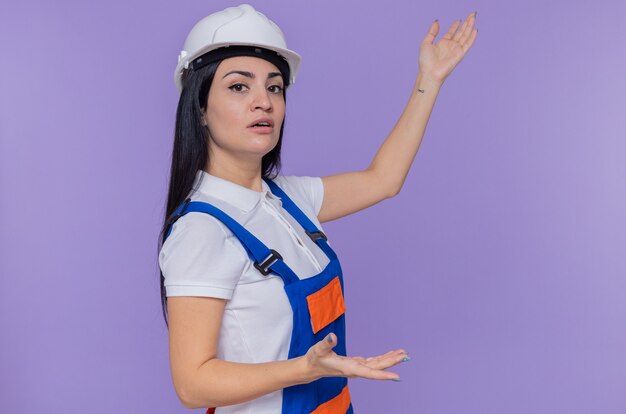 Giovane donna del costruttore in uniforme da costruzione e casco di sicurezza guardando la parte anteriore con il viso serio che presenta qualcosa con le braccia in piedi sul muro viola