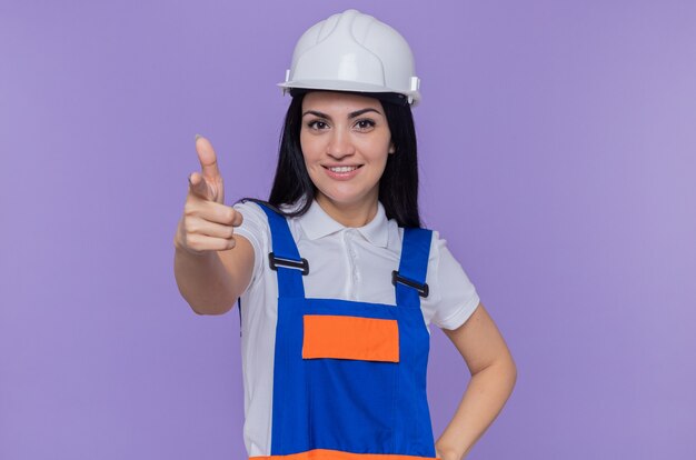 Giovane donna del costruttore in uniforme da costruzione e casco di sicurezza sorridente che punta fiducioso con il dito indice davanti in piedi sopra la parete viola