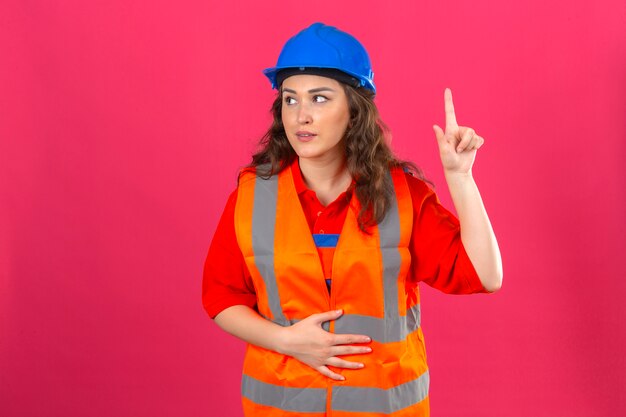 Giovane donna del costruttore in uniforme della costruzione e casco di sicurezza che distolgono lo sguardo indicando su con il dito indice sopra la parete rosa isolata