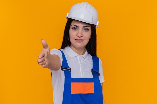 Giovane donna del costruttore in uniforme della costruzione e casco di sicurezza che esamina fronte sorridente fiducioso che offre il gesto di saluto della mano che sta sopra la parete arancione