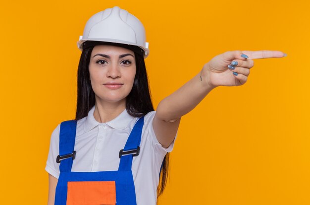 Giovane donna del costruttore in uniforme della costruzione e casco di sicurezza che esamina il puntamento sicuro sorridente anteriore con il dito indice al lato che sta sopra la parete arancione