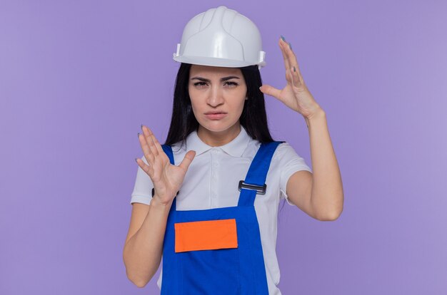 Giovane donna del costruttore in uniforme della costruzione e casco di sicurezza che esamina la parte anteriore concentrandosi su un compito che fa gesto di dimensione con le mani che stanno sopra la parete viola