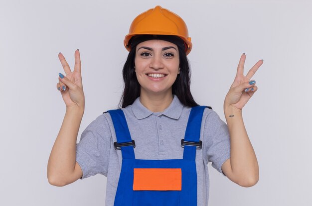 Giovane donna del costruttore in uniforme della costruzione e casco di sicurezza che esamina sorridere felice e positivo anteriore che mostra il segno di v che sta sopra il muro bianco