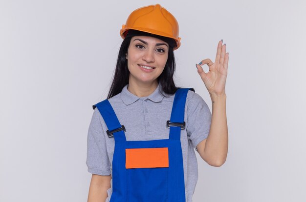 Giovane donna del costruttore in uniforme della costruzione e casco di sicurezza che esamina sorridere felice e positivo anteriore che mostra il segno giusto che sta sopra il muro bianco