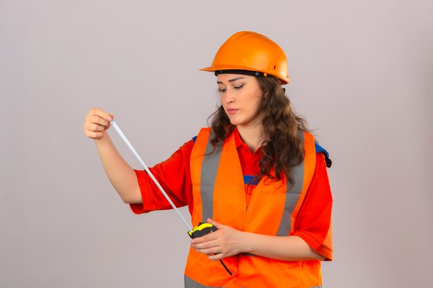 Giovane donna del costruttore in uniforme della costruzione e casco di sicurezza che esaminano nastro di misura con il fronte serio sopra la parete bianca isolata