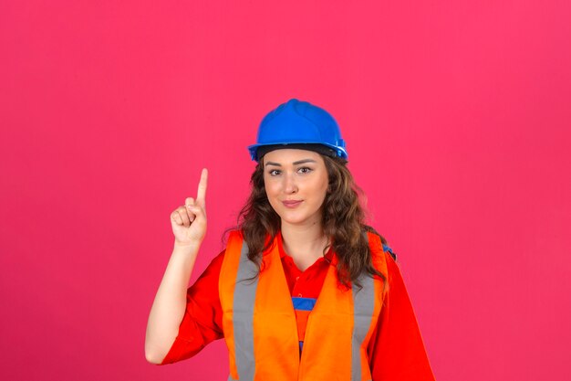 Giovane donna del costruttore in uniforme della costruzione e casco di sicurezza che indicano su con il nuovo concetto di idea del dito sopra la parete rosa isolata