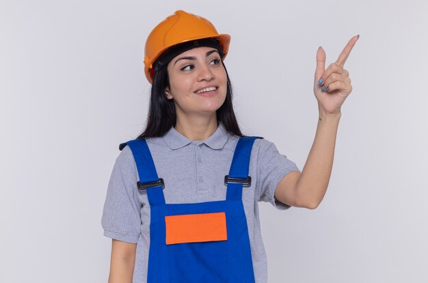 Giovane donna del costruttore in uniforme della costruzione e casco di sicurezza che osserva da parte con il sorriso sul fronte felice che indica con il dito indice a qualcosa che sta sopra il muro bianco