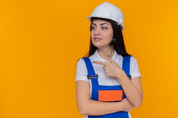 Giovane donna del costruttore in uniforme della costruzione e casco di sicurezza che osserva da parte