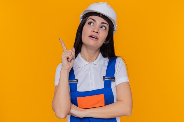 Giovane donna del costruttore in uniforme della costruzione e casco di sicurezza che osserva in su con il sorriso sul pensiero astuto del fronte che mostra il dito indice che ha grande idea che sta sopra la parete arancione