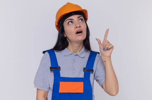 Giovane donna del costruttore in uniforme della costruzione e casco di sicurezza che osserva in su sorpreso che indica con il dito indice a qualcosa che si leva in piedi sopra la parete bianca