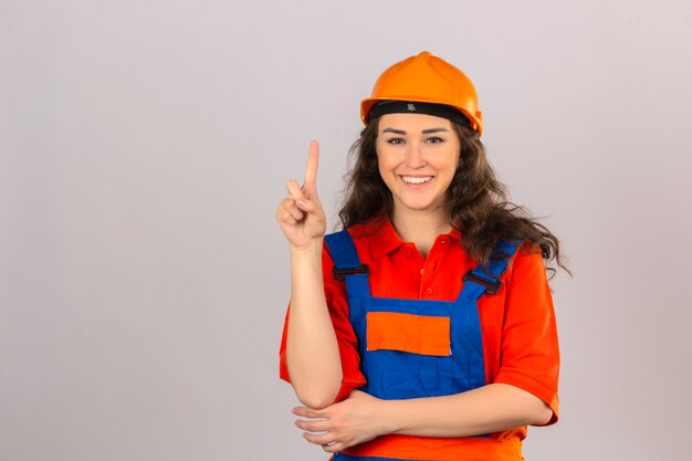 Giovane donna del costruttore in uniforme della costruzione e casco di sicurezza che sembrano dito indicante sicuro su sopra la parete bianca isolata