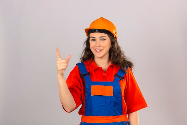 Giovane donna del costruttore in uniforme della costruzione e casco di sicurezza che sembrano dito indicante sicuro su sopra la parete bianca isolata
