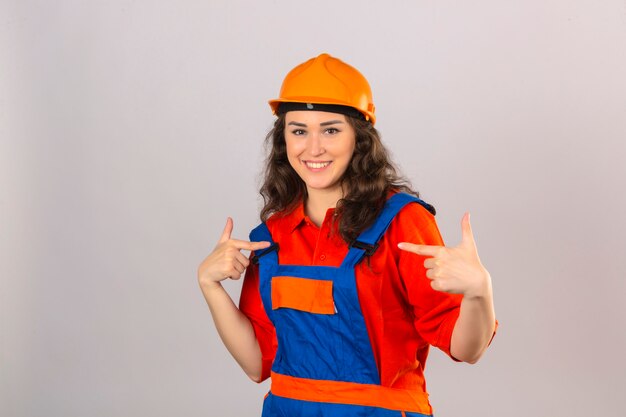 Giovane donna del costruttore in uniforme della costruzione e casco di sicurezza che sembrano indicare sicuro se stessa sopra la parete bianca isolata