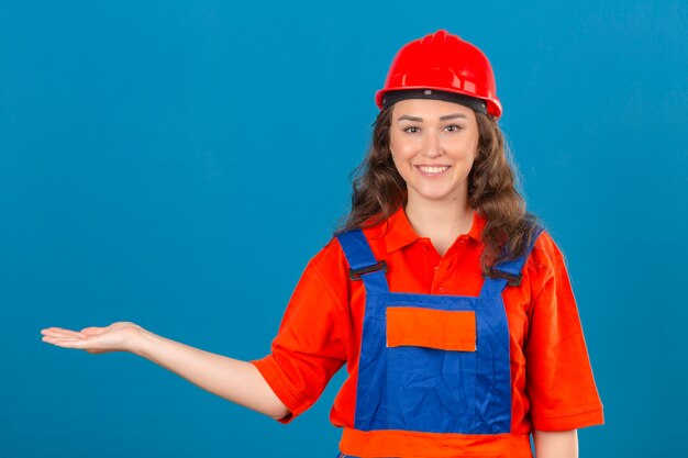 Giovane donna del costruttore in uniforme della costruzione e casco di sicurezza che sorridono presentazione allegra e che indicano con il palmo della mano che esamina la macchina fotografica sopra la parete blu isolata