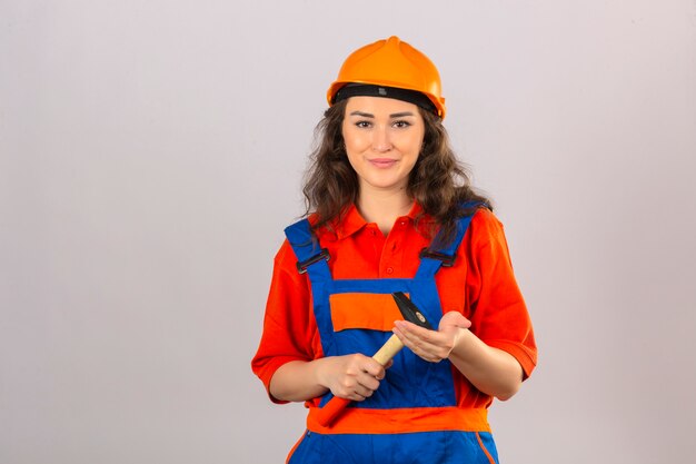 Giovane donna del costruttore in uniforme della costruzione e casco di sicurezza che stanno con il martello in mani che sorridono sopra la parete bianca isolata