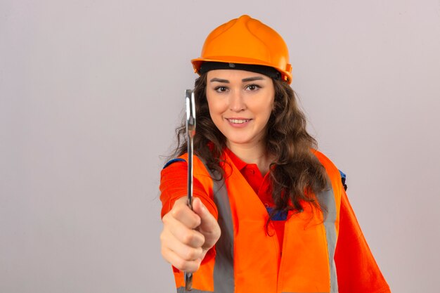 Giovane donna del costruttore in uniforme della costruzione e casco di sicurezza che stanno con la chiave che lo mostra alla macchina fotografica che sorride amichevole sopra la parete bianca isolata