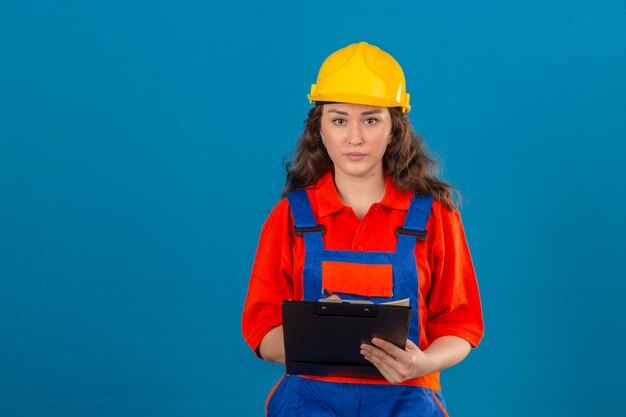 Giovane donna del costruttore in uniforme della costruzione e casco di sicurezza che stanno con la lavagna per appunti in mani con il fronte serio sopra la parete blu isolata