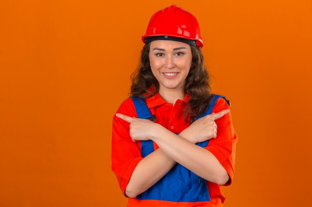 Giovane donna del costruttore in uniforme della costruzione e casco di sicurezza che stanno con le armi attraversate e che indicano con i dito indice agli altri lati che sorridono sopra la parete arancio isolata