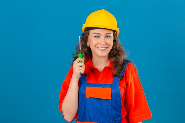 Giovane donna del costruttore in uniforme della costruzione e casco di sicurezza che stanno con sorridere amichevole del cacciavite sopra la parete blu isolata