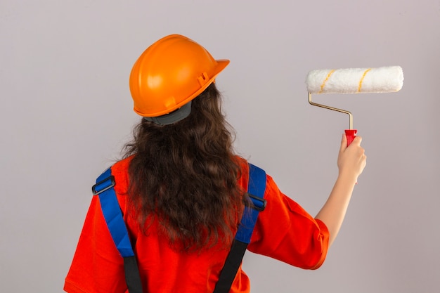 Giovane donna del costruttore in uniforme della costruzione e casco di sicurezza che stanno indietro con il rullo di pittura sopra la parete bianca isolata