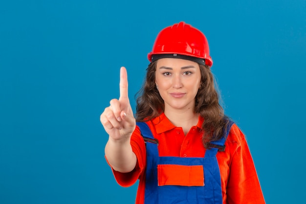 Giovane donna del costruttore in uniforme della costruzione e casco di sicurezza con il sorriso sul fronte che mostra numero uno con il dito indice sopra la parete blu isolata