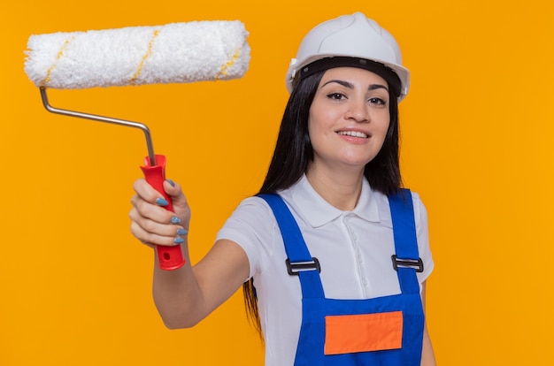 Giovane donna del costruttore in uniforme di costruzione e casco di sicurezza che tiene il rullo di vernice guardando davanti sorridente in piedi sicuro sopra la parete arancione