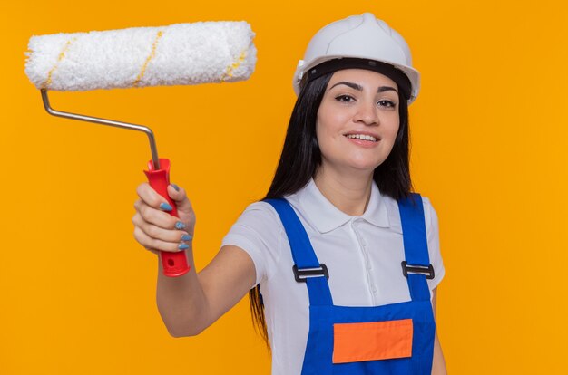 Giovane donna del costruttore in uniforme di costruzione e casco di sicurezza che tiene il rullo di vernice guardando davanti sorridente in piedi sicuro sopra la parete arancione