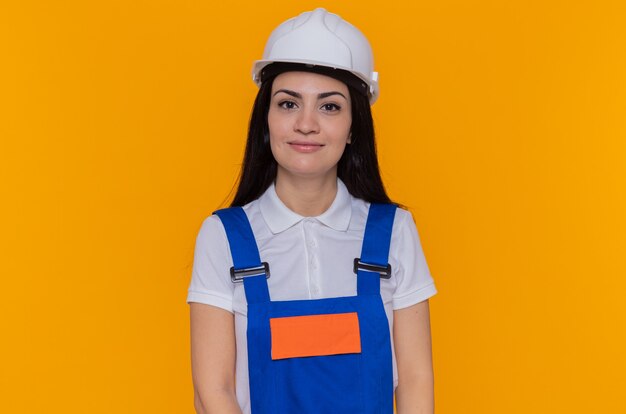 Giovane donna del costruttore in uniforme di costruzione e casco di sicurezza guardando davanti sorridente fiducioso in piedi sopra la parete arancione