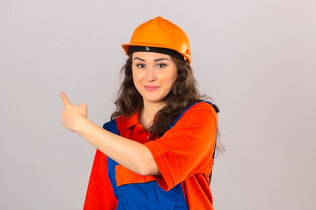 Giovane donna del costruttore nell'uniforme della costruzione e nell'indicare sorridente del casco di sicurezza con la mano ed il dito il lato sopra la parete bianca isolata