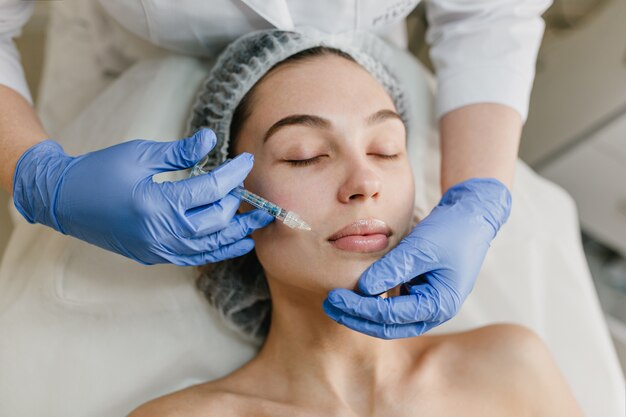 Giovane donna del ritratto durante le procedure di cosmetologia nel salone di bellezza. Iniezione, botox, mani in bagliori blu, assistenza sanitaria, terapia, labbra, bellezza