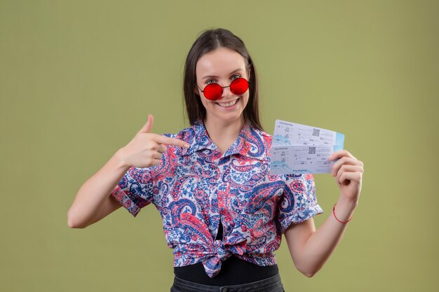 Giovane donna del viaggiatore che indossa gli occhiali da sole rossi che tengono i biglietti che indicano con il dito loro che sorridono con il fronte felice sopra la parete verde