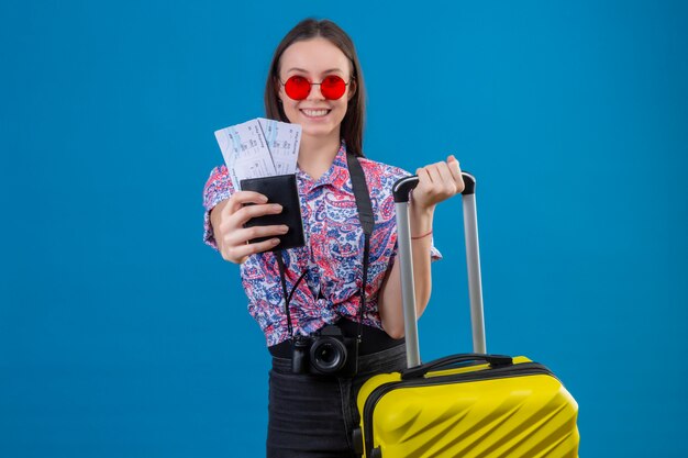 Giovane donna del viaggiatore che indossa gli occhiali da sole rossi con la valigia gialla che mostra passaporto e biglietti che sorridono allegramente con il fronte felice sopra la parete blu