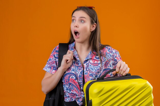 Giovane donna del viaggiatore che indossa gli occhiali da sole rossi sulla testa con la valigia della tenuta dello zaino che guarda da parte sorpresa