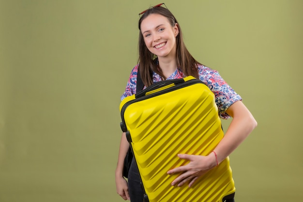 Giovane donna del viaggiatore che indossa gli occhiali da sole rossi sulla testa con la valigia della tenuta dello zaino che sorride allegramente con il fronte felice sopra la parete verde isolata