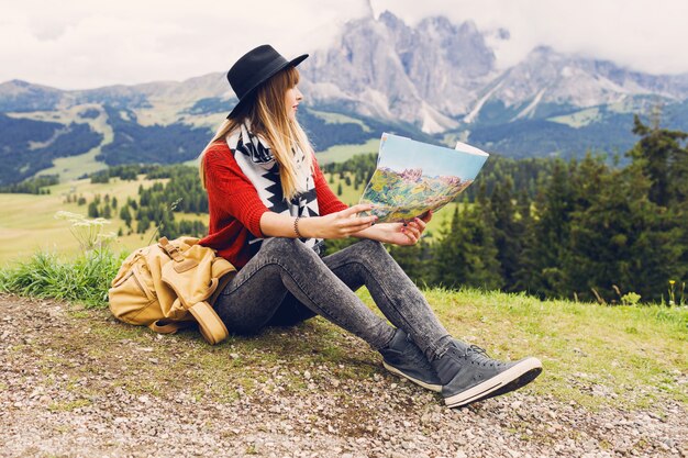 Giovane donna del viaggiatore con lo zaino e il cappello che si siedono sull'erba e che cercano la giusta direzione sulla mappa
