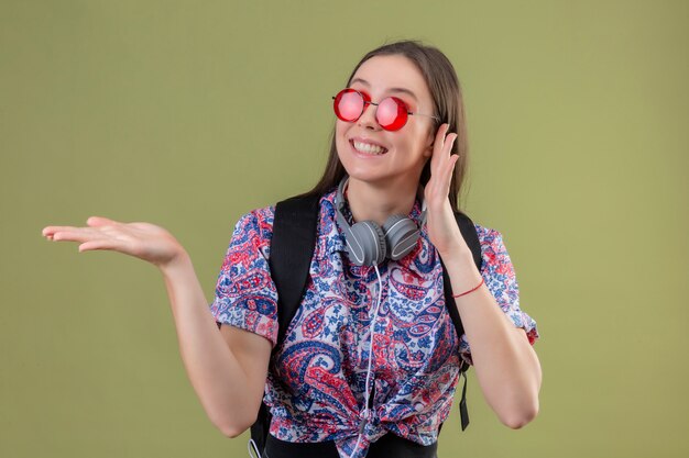 Giovane donna del viaggiatore con lo zaino e le cuffie che indossano gli occhiali da sole rossi che sorridono allegramente che presentano con il braccio della mano che controlla parete verde