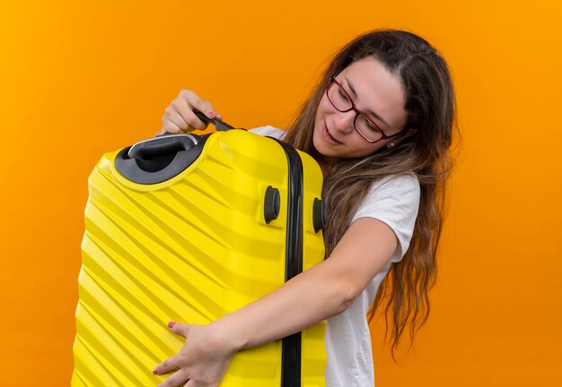 Giovane donna del viaggiatore in maglietta bianca che abbraccia la valigia in piedi felice e positivo sopra la parete arancione