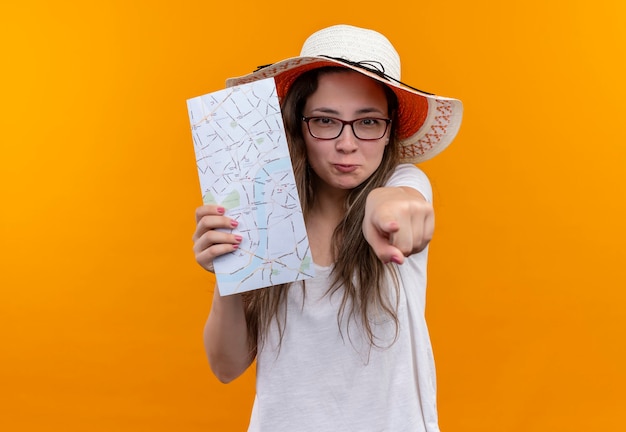 Giovane donna del viaggiatore in maglietta bianca che indossa cappello estivo che tiene la mappa che indica con il dito indice nella parte anteriore