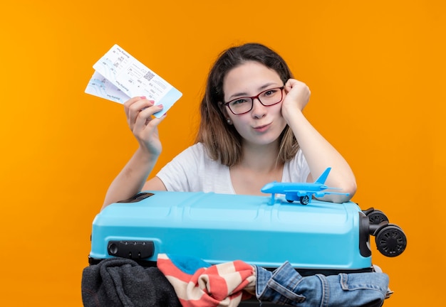 Giovane donna del viaggiatore in maglietta bianca che tiene la valigia piena di vestiti e biglietti aerei appoggiato la testa a portata di mano guardando sorridente in piedi