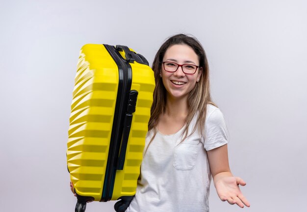 Giovane donna del viaggiatore in t-shirt bianca che tiene la valigia guardando sorridente allegramente alzando il braccio in piedi sopra il muro bianco