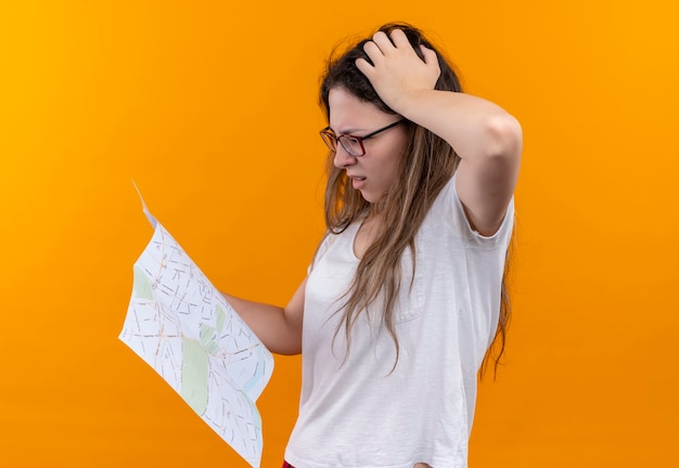 Giovane donna del viaggiatore in t-shirt bianca che tiene mappa guardandolo con espressione pensierosa sul viso che sembra confuso in piedi sopra la parete arancione