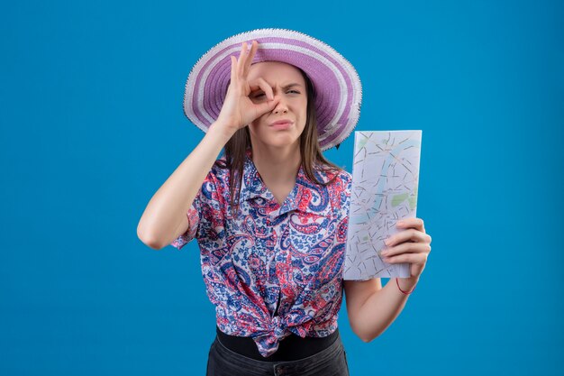 Giovane donna del viaggiatore nella mappa della tenuta del cappello di estate che fa segno giusto che guarda attraverso questo segno con l'espressione sospettosa sopra la parete blu