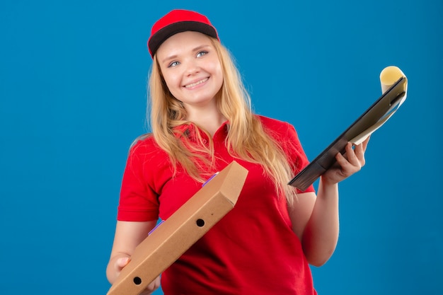Giovane donna delle consegne che indossa la maglietta polo rossa e cappuccio in piedi con la scatola della pizza e appunti con il sorriso sul viso su sfondo blu isolato